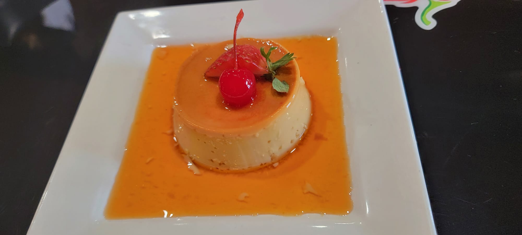 Flan casero in caramel sauce topped with a cherry, strawberry and mint on a white plate