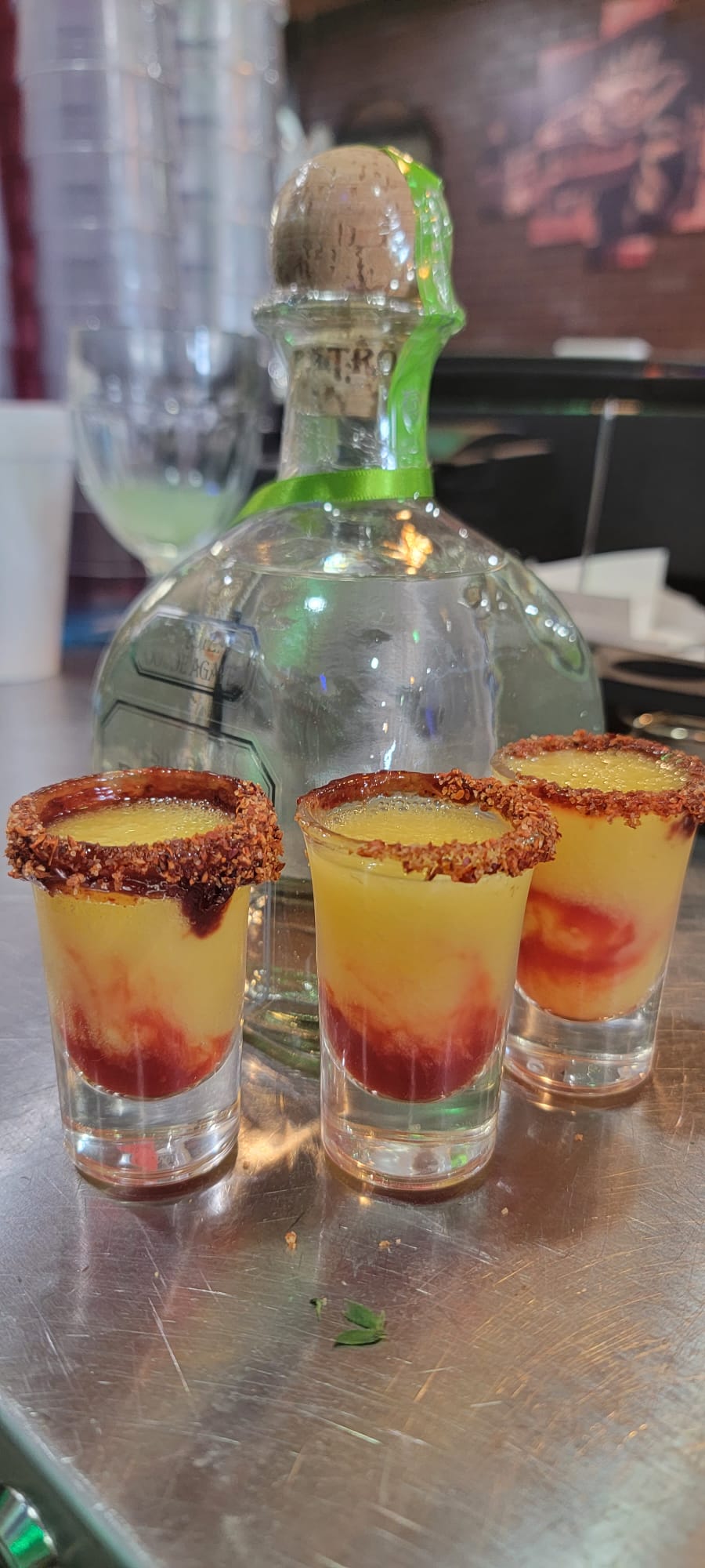 Three mangonada shots with chili-chamoy rims layered with mango and chamoy beside a tequila bottle