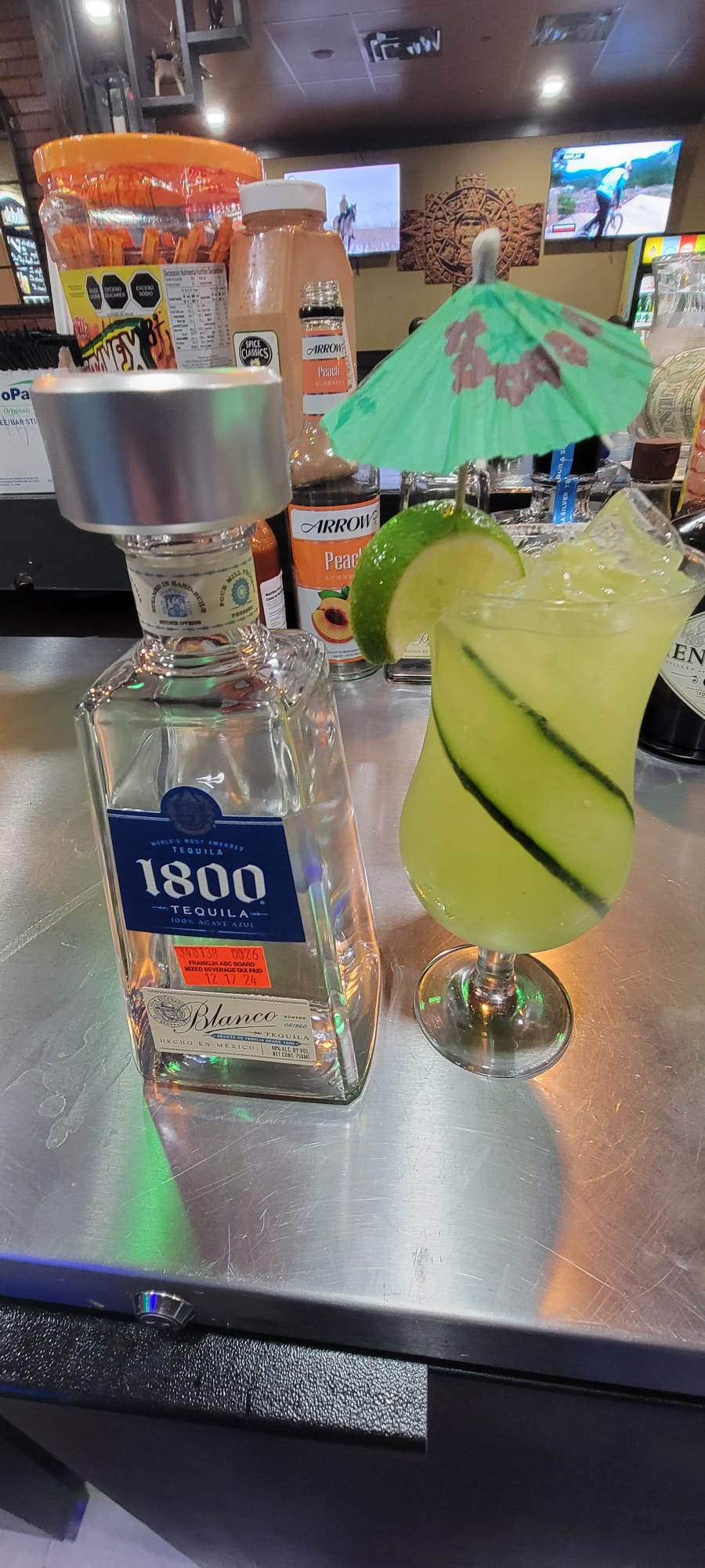 Cucumber margarita with lime and paper umbrella beside a bottle of blanco tequila