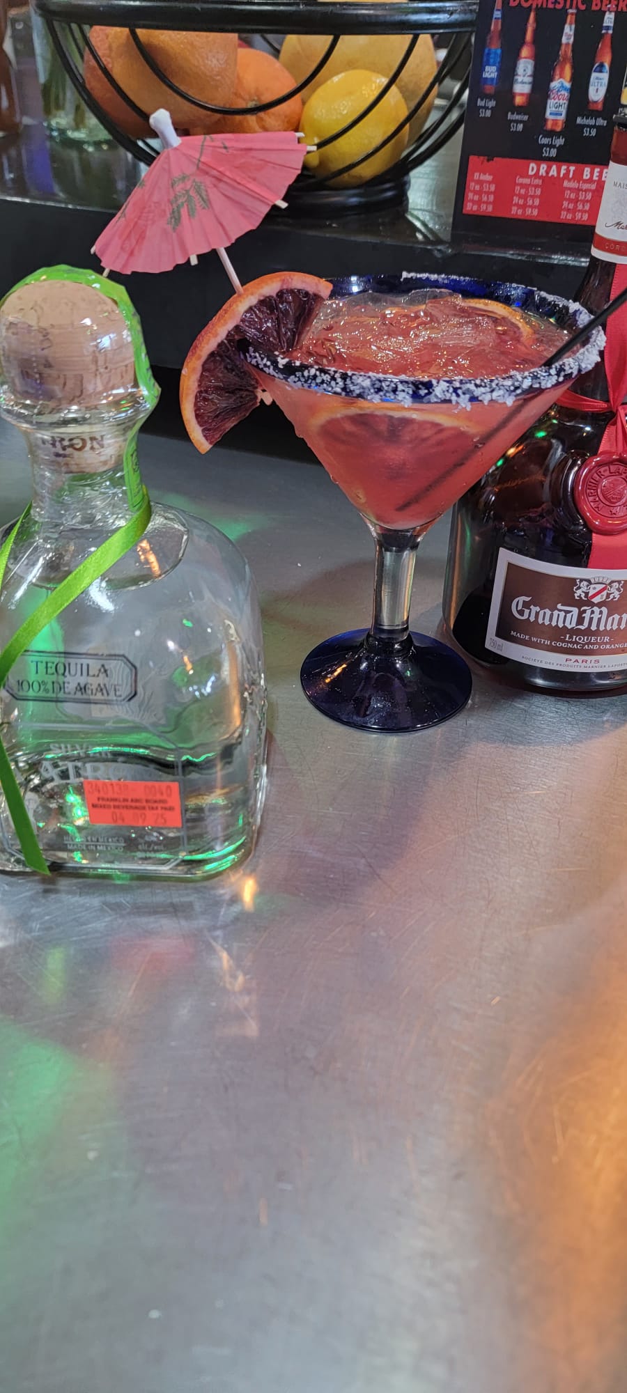 Blood orange margarita in a blue-rimmed martini glass with salt rim, orange wheel and pink paper umbrella